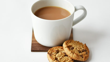 White ceramic mug of a light-brown drink on a square wooden coaster beside two chocolate-chip cookies on a white tabletop