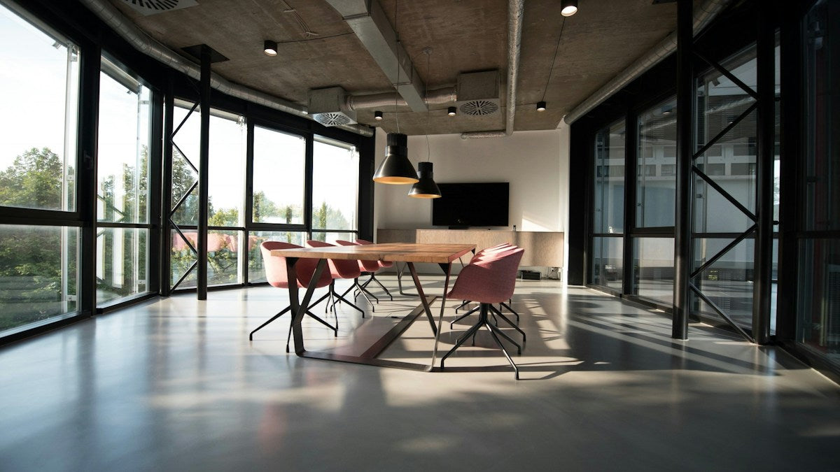 Sunlit meeting room with a wooden table, pink chairs, large windows, and greenery outside