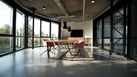 Sunlit meeting room with a wooden table, pink chairs, large windows, and greenery outside