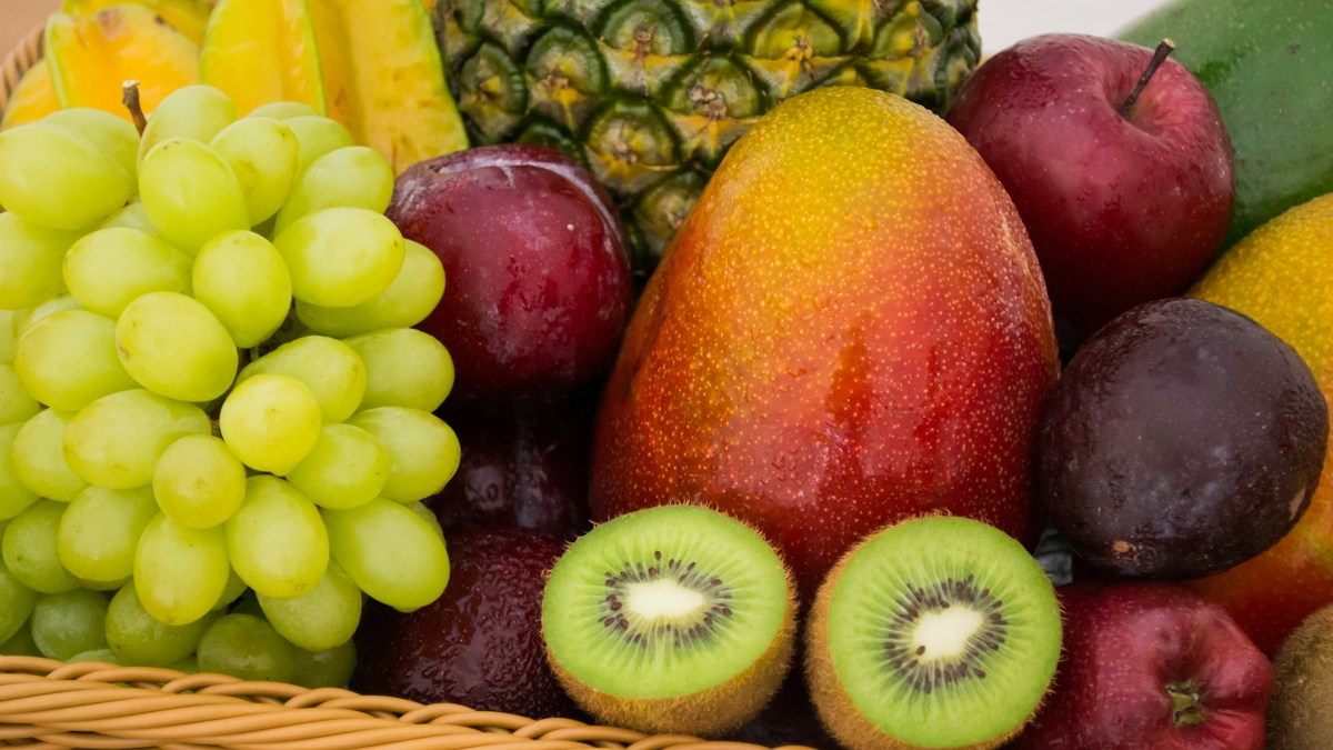 Basket of fresh fruit with green grapes, mango, kiwi halves, dark plums, a red apple, pineapple, and starfruit