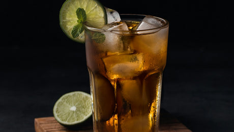 Faceted glass of an iced amber drink with a lime wheel and mint sprig on a wooden surface against a dark background