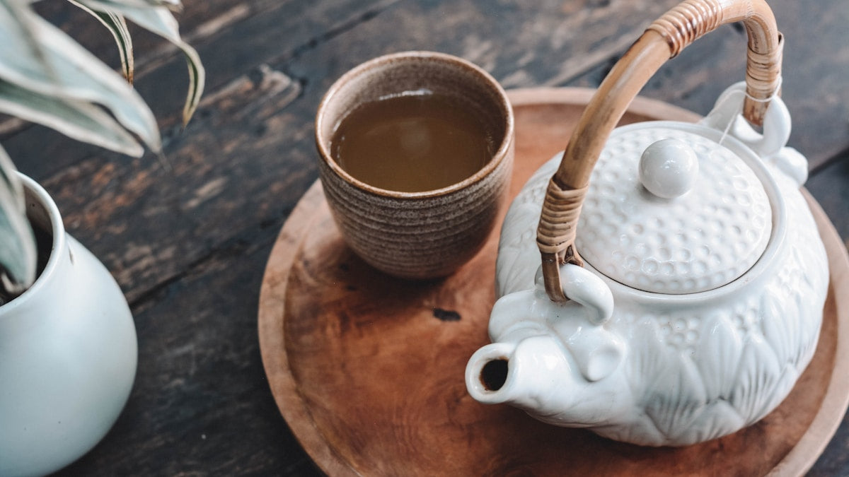 Amber tea in a brown ceramic cup beside a white teapot on a wooden tray