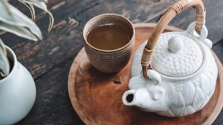 Amber tea in a brown ceramic cup beside a white teapot on a wooden tray