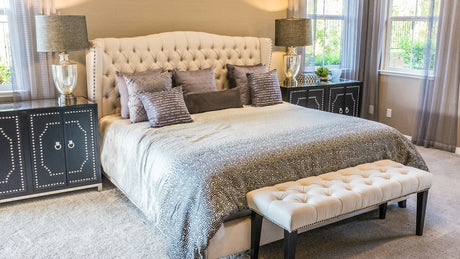 Bright bedroom with a cream tufted headboard, layered gray bedding, matching bedside lamps, and natural window light